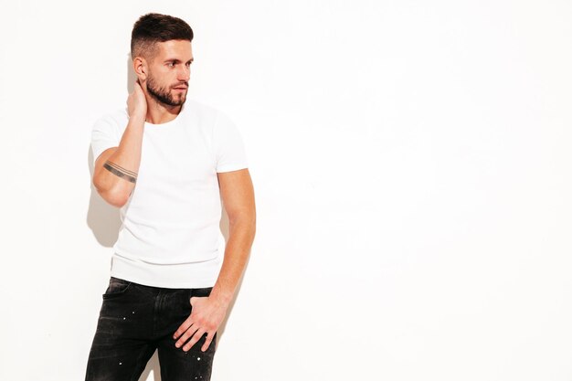 Retrato de guapo confiado con estilo hipster lambersexual modelMan vestido con camiseta blanca y jeans Moda masculina posando cerca de la pared en el estudio aislado