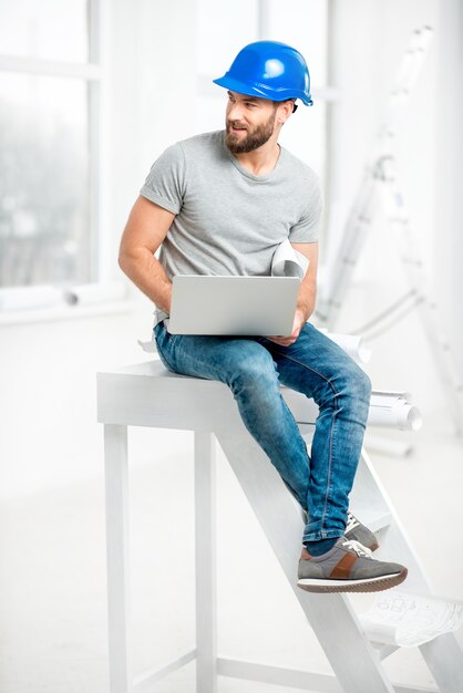 Retrato de un guapo constructor, capataz o reparador en el casco sentado con una computadora portátil en el interior blanco