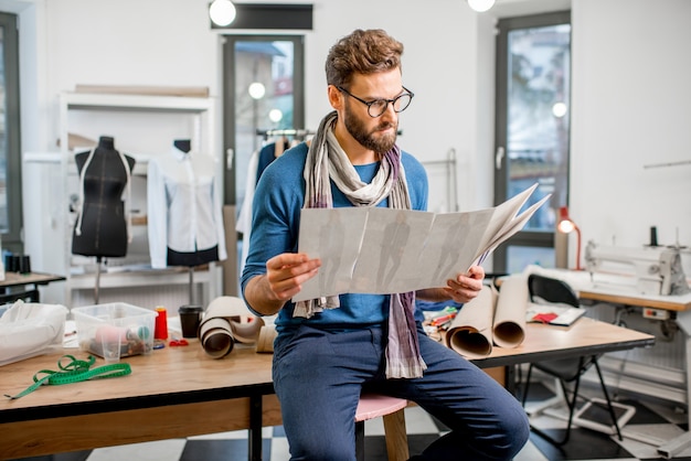 Retrato de un guapo diseñador de moda sentado con bocetos de ropa en el estudio lleno de ropa y herramientas de confección