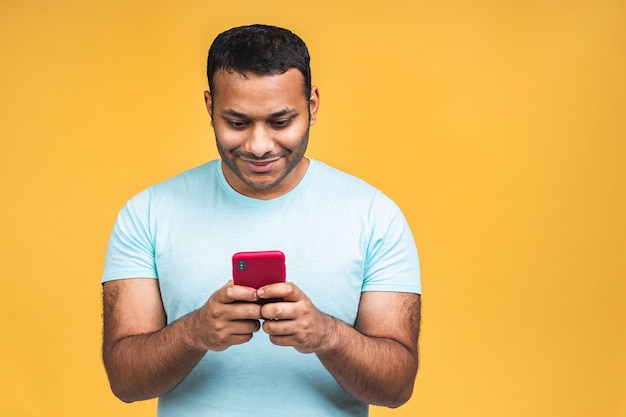 Retrato de guapo, emocionado, alegre, alegre, indio, afroamericano, llevando, casual, envío, y, recibiendo, mensajes, a, su, amante, aislado, encima, fondo amarillo. Usando el teléfono.