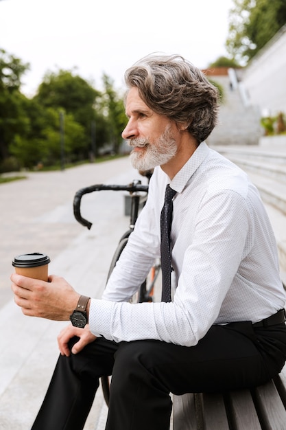 Retrato de guapo empresario maduro en ropa formal bebiendo café para llevar mientras está sentado en un banco con bicicleta al aire libre