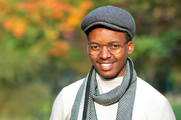 Retrato de guapo feliz positivo alegre chico negro, joven afroamericano étnico afroamericano está caminando, sonriendo en el parque de otoño dorado en el clima soleado de otoño en gafas, sombrero y bufanda. Felicidad