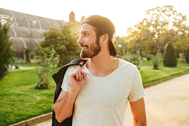 Retrato de guapo hombre barbudo de 30 años con gorra y camiseta blanca con chaqueta de cuero en el hombro durante la caminata en el parque verde