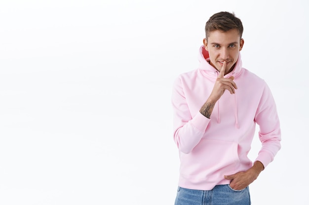 Retrato de guapo joven rubio descarado en sudadera con capucha rosa, sonriendo escondiendo secreto, pidiendo silencio