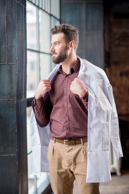 Retrato de guapo médico seguro con bata médica de pie cerca de la ventana en la moderna clínica u oficina interior oscuro