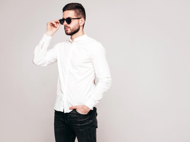 Retrato de guapo modelo confiado Hombre elegante sexy vestido con camisa blanca y jeans Hombre hipster de moda posando en estudio sobre fondo gris En gafas de sol Aislado