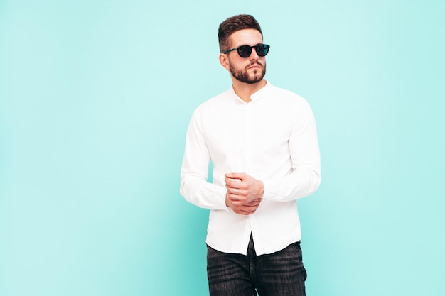 Retrato de guapo modelo confiado Hombre elegante sexy vestido con camisa blanca y jeans Hombre hipster de moda posando junto a la pared azul en el estudio Aislado en gafas de sol