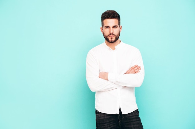 Retrato de guapo modelo confiado Hombre elegante sexy vestido con camisa blanca y jeans Hombre hipster de moda posando junto a la pared azul en el estudio Brazos cruzados Aislado