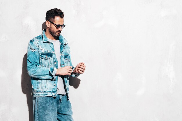 Retrato de guapo modelo lambersexual con estilo hipster seguro Hombre sexy vestido con chaqueta de jeans Hombre de moda aislado en la pared gris en el estudio En gafas de sol