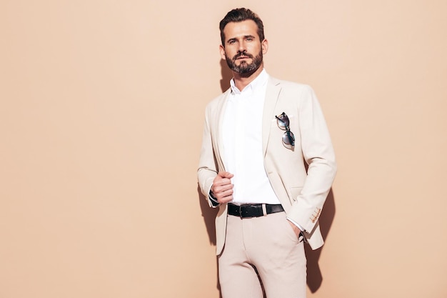 Retrato de guapo modelo lambersexual con estilo hipster sonriente Hombre moderno sexy vestido con elegante traje blanco Hombre de moda con peinado rizado posando en el estudio cerca de la pared beige