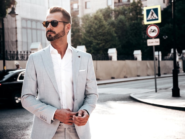 Retrato de guapo modelo lambersexual hipster con estilo seguro Hombre moderno sexy vestido con elegante traje blanco Hombre de moda posando en el fondo de la calle en la ciudad de Europa al atardecer Con gafas de sol