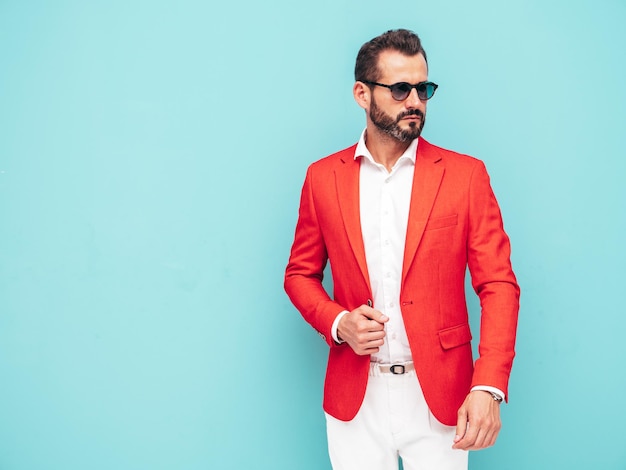 Retrato de guapo modelo lambersexual hipster con estilo seguro Hombre moderno sexy vestido con elegante traje rojo Hombre de moda posando en el estudio cerca de la pared azul con gafas de sol