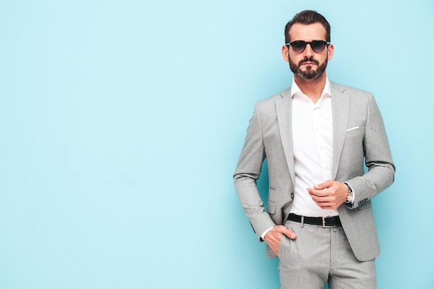 Retrato de guapo modelo lambersexual hipster con estilo seguro Hombre moderno sexy vestido con traje elegante Hombre de moda posando en el estudio cerca de la pared azul con gafas de sol