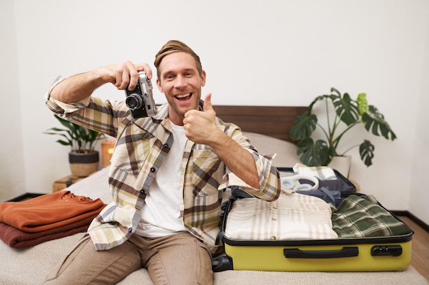 Retrato de un guapo modelo masculino turista con una cámara sentado cerca de una maleta abierta con ropa