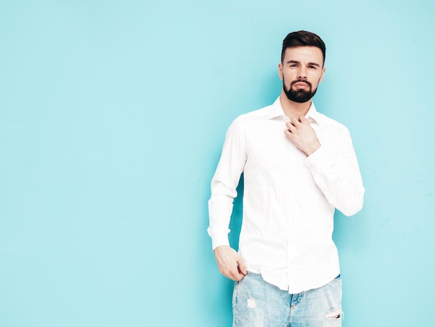 Retrato de guapo modelo sonriente Hombre elegante sexy vestido con camisa blanca y jeans Hombre hipster de moda posando junto a la pared azul en el estudio Aislado