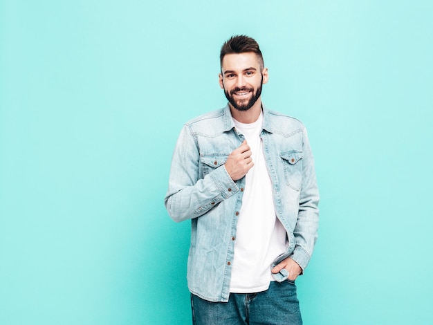 Retrato de guapo sonriente con estilo hipster lambersexual modelMan vestido con chaqueta y jeans Moda masculina posando cerca de la pared azul en el estudio aislado