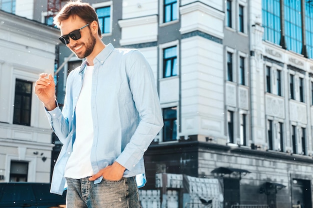 Retrato de guapo sonriente con estilo hipster lambersexual modelModern hombre vestido con camisa azul Fashion male posando cerca de rascacielos en el fondo de la calle en gafas de sol al aire libre al atardecer