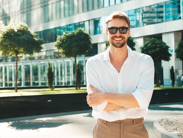 Retrato de guapo sonriente con estilo hipster lambersexual modelModern hombre vestido con camisa blanca Hombre de moda posando en el fondo de la calle cerca de rascacielos en gafas de sol Al aire libre al atardecer