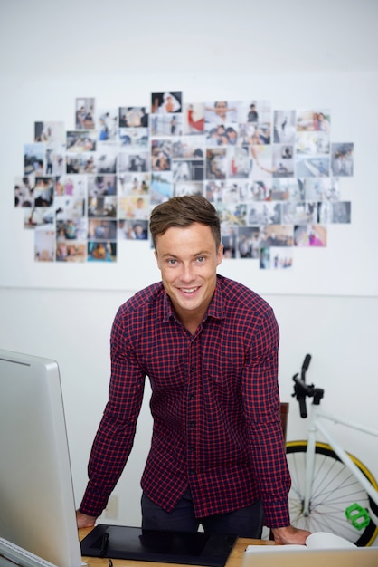 Retrato de guapo sonriente joven diseñador gráfico en camisa a cuadros apoyado en el escritorio con computadora y tableta gráfica y sonriendo a la cámara