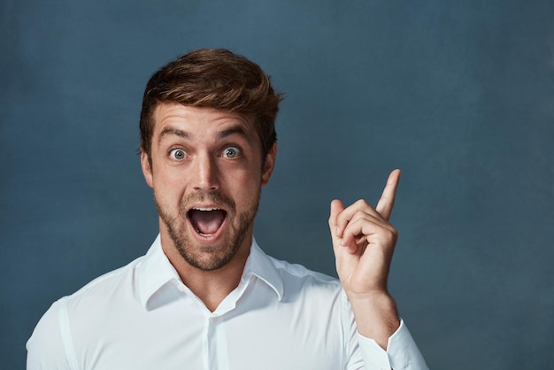 Retrato guau e idea con un hombre señalando en el estudio sobre un fondo azul durante un momento de bombilla Sorpresa facial y eureka con un joven emocionado que parece sorprendido o asombrado por las buenas noticias