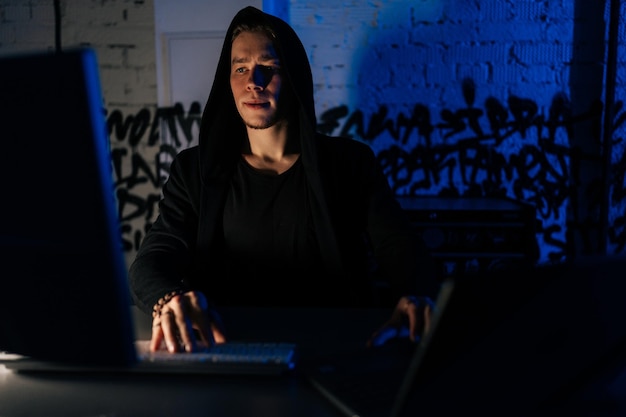 Retrato de un hacker oculto con sudadera con capucha dedicado a piratear sistemas de seguridad sentado en un sótano oscuro con luces de neón azules