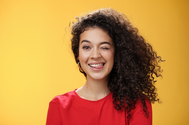 Retrato de hermana divertida y fresca con cabello rizado guiñando un ojo juguetonamente divirtiéndose y tonteando mostrando la lengua mientras juega con hermanos adorando pasar tiempo con niños sobre fondo amarillo.