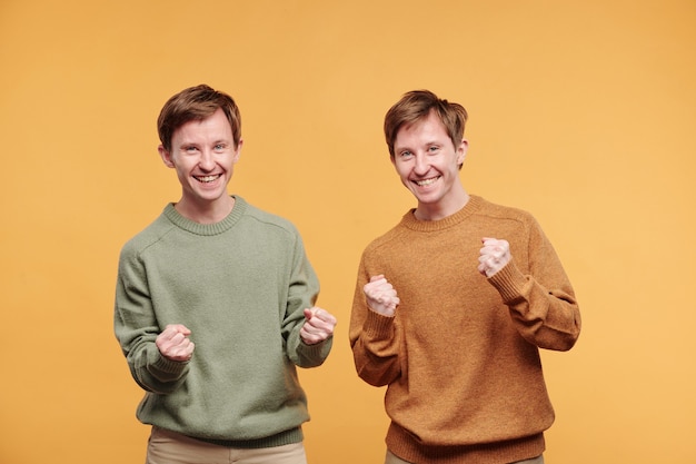 Retrato de hermanos gemelos emocionados emocionados en suéteres casuales haciendo gestos de sí contra el fondo naranja