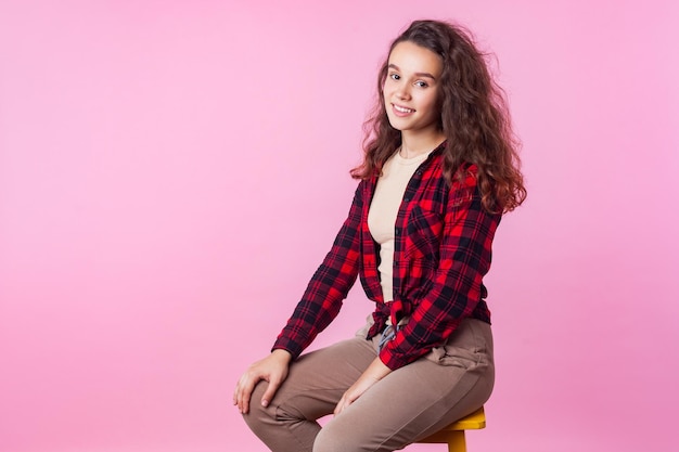Foto retrato de una hermosa adolescente de moda con cabello largo y rizado de morena en camisa a cuadros sentada relajada en una silla y mirando a la cámara con una linda sonrisa en un estudio interior aislado en un fondo rosa