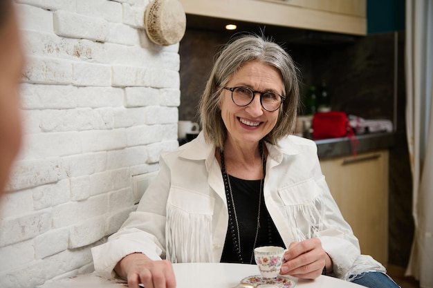 Retrato de una hermosa anciana caucásica con ropa elegante sentada en la mesa y disfrutando de un descanso para tomar café en su acogedor estudio