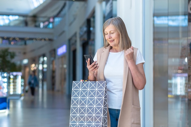 Retrato de una hermosa anciana de compras en el centro comercial Ella usa el teléfono hace compras en línea ganó un regalo se regocija Sostiene bolsas de papel en sus manos Ella sonríe feliz con sus compras