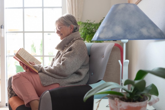Retrato de una hermosa anciana relajada sentada en su casa en un sillón leyendo un libro sobre la vieja generación y el concepto de jubilación