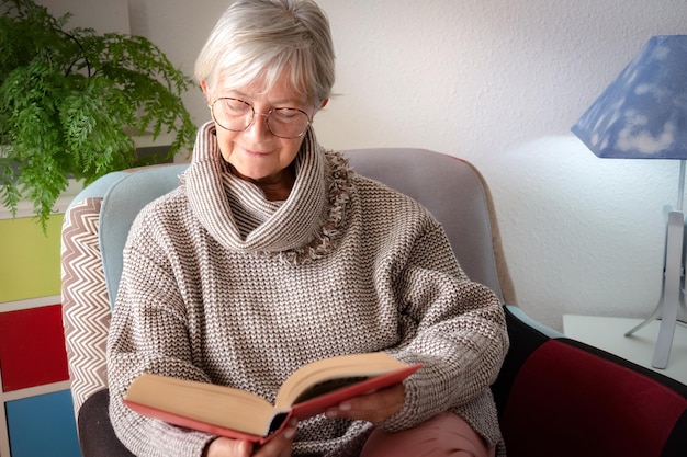 Retrato de una hermosa anciana relajada sentada en su casa en un sillón leyendo un libro sobre la vieja generación y el concepto de jubilación