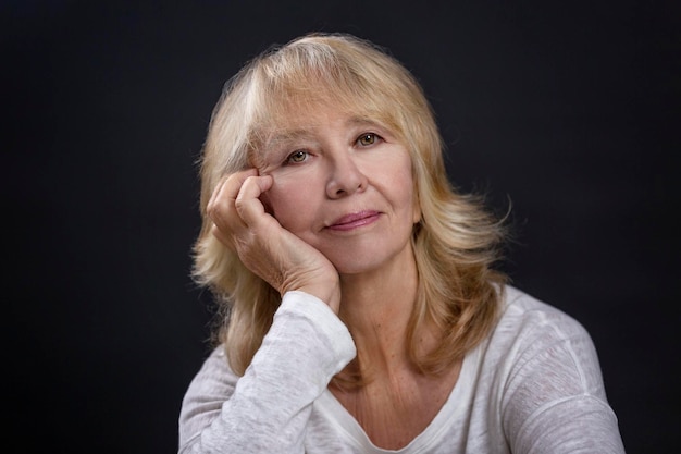 Foto retrato de una hermosa anciana una rubia sonriente se sienta con la mano apoyando su mejilla feliz madurez closeup fondo negro