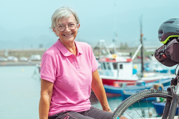 Retrato de una hermosa anciana sentada en un banco en el puerto marítimo cerca de su bicicleta eléctrica Concepto de libertad y vacaciones