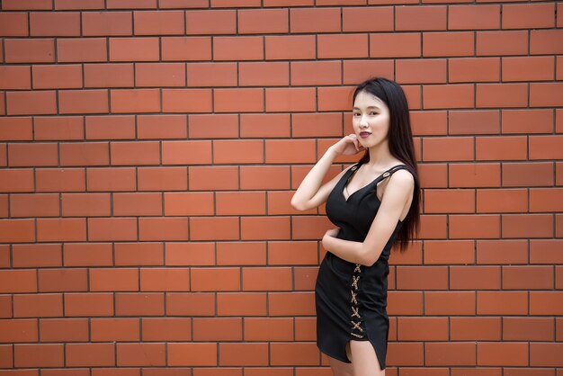 Retrato de una hermosa chica asiática elegante que usa un vestido negro para tomar una foto en una pared de ladrillos Estilo de vida de los adolescentes de Tailandia Concepto feliz de mujer moderna