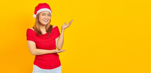 Retrato de una hermosa chica asiática con sombrero de navidad apuntando al espacio vacío con la palma aislada en el fondo amarillo