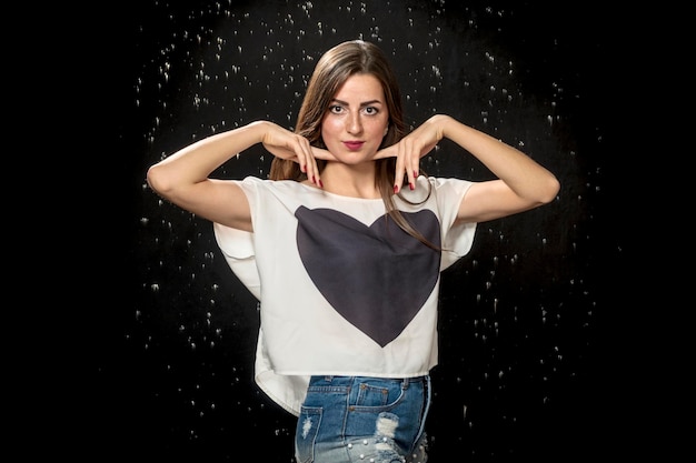 retrato de una hermosa chica en una camiseta con corazón y dedos en el cuello sobre el fondo de gotas de agua sobre un fondo negro.