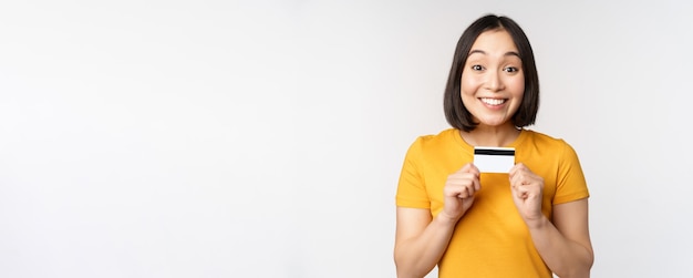 Retrato de una hermosa chica coreana sosteniendo una tarjeta de crédito recomendando el servicio bancario de pie en una camiseta amarilla sobre fondo blanco