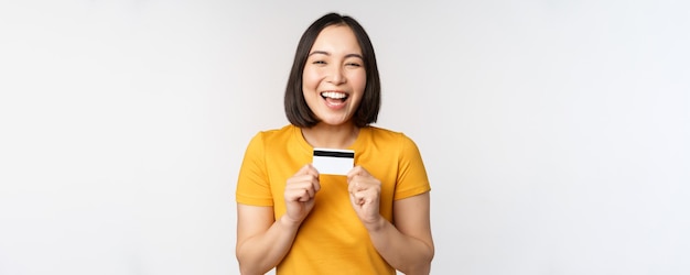 Retrato de una hermosa chica coreana sosteniendo una tarjeta de crédito recomendando el servicio bancario de pie en una camiseta amarilla sobre fondo blanco