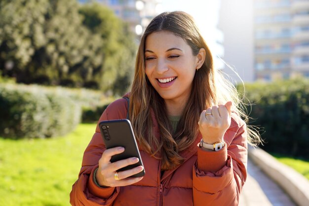 Retrato de una hermosa chica emocionada leyendo buenas noticias en el teléfono móvil al aire libre