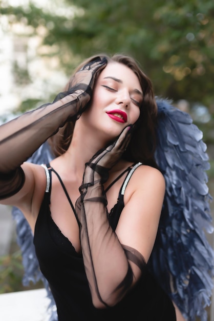 Retrato de una hermosa chica con labios rojos en un traje negro, guantes y alas azules Posando disfrutando tocándose la cara con las manos