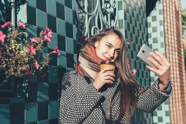 Retrato de una hermosa chica morena con bebida para llevar en la calle. bufanda y abrigo de otoño en la ciudad. Impresionante vista de la dama de negocios con una taza de café caminando por la calle de la ciudad.