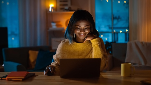 Retrato de una hermosa chica negra que usa la computadora mientras está sentada en su escritorio en casa, lleva un suéter cálido por la noche, una mujer creativa trabaja en una computadora en su acogedora sala de estar.