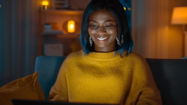 Retrato de una hermosa chica negra que usa la computadora mientras se relaja en un sofá en casa Su cara está iluminada por una pantalla por la noche Una mujer creativa trabaja en una computadora en su acogedora sala de estar