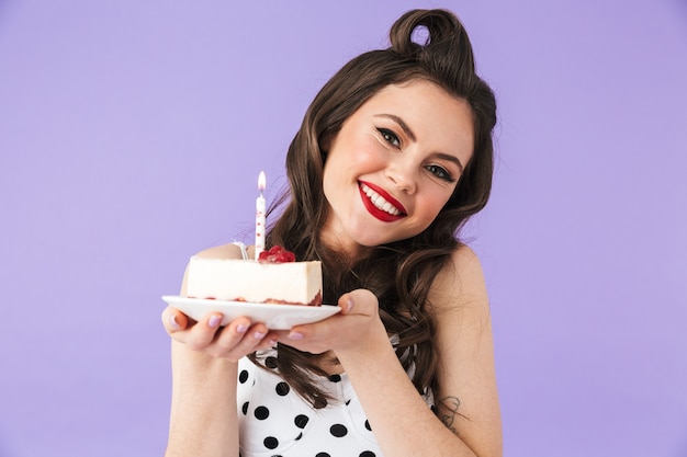 Retrato de una hermosa chica pin-up con maquillaje brillante que se encuentran aisladas sobre la pared violeta, sosteniendo un plato con un trozo de tarta