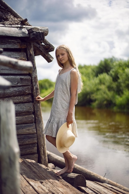 Retrato de una hermosa chica de pueblo rubia con el pelo largo en un vestido y sombrero de pie junto a una antigua casa de madera en verano uno del río