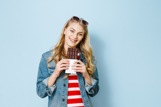 Un retrato de una hermosa chica rubia comiendo chocolate está emocionado sobre un fondo azul.