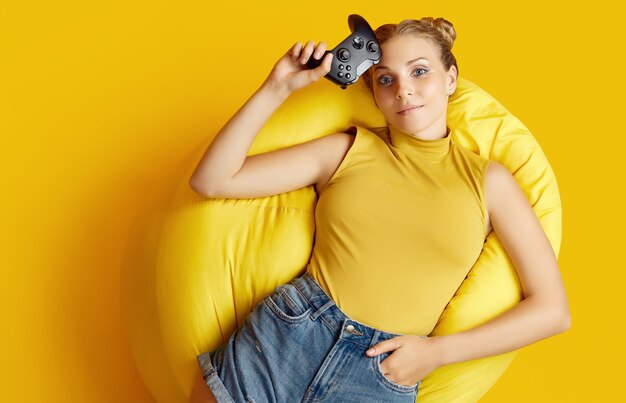 Retrato de hermosa chica rubia feliz gamer con joystick acostado en una silla de saco sobre superficie amarilla