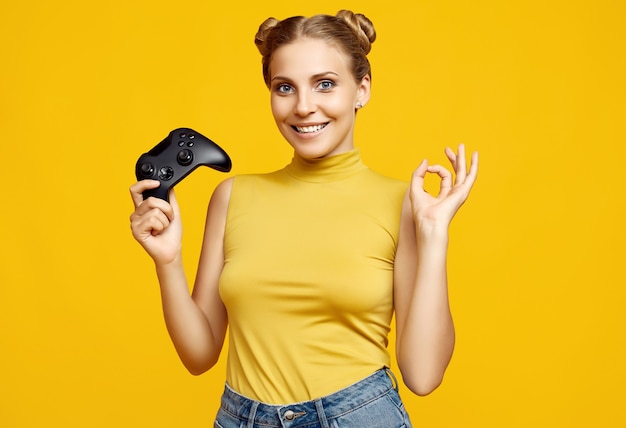 Foto retrato de hermosa chica rubia feliz gamer jugando videojuegos con joystick en pared amarilla en estudio