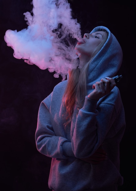 Foto retrato de hermosa chica rubia en sudadera con capucha gris fuma vape aislado sobre fondo negro de estudio, nube de humo de vapor, mini cachimba